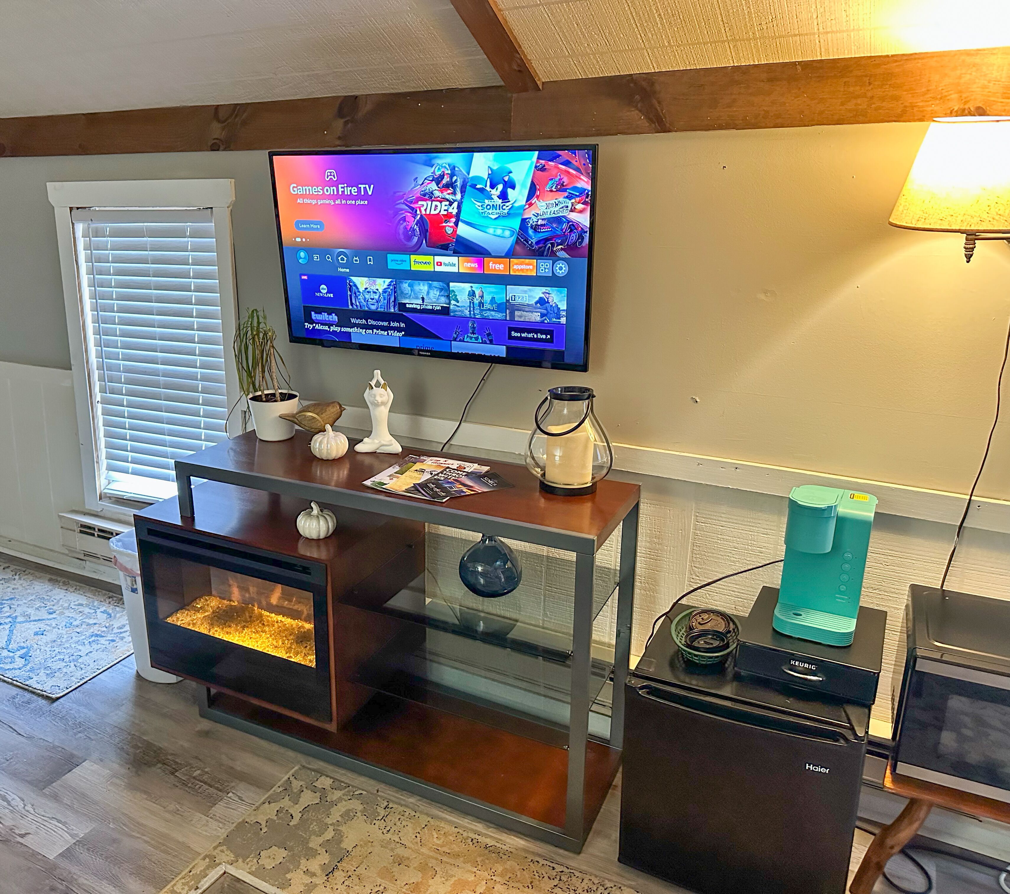 Una sala de estar con un televisor montado en la pared mostrando varios canales de videojuegos, un mueble de entretenimiento de madera con puerta de vidrio, una pequeña mesa de centro y una cafetera Keurig sobre un electrodoméstico Haier.