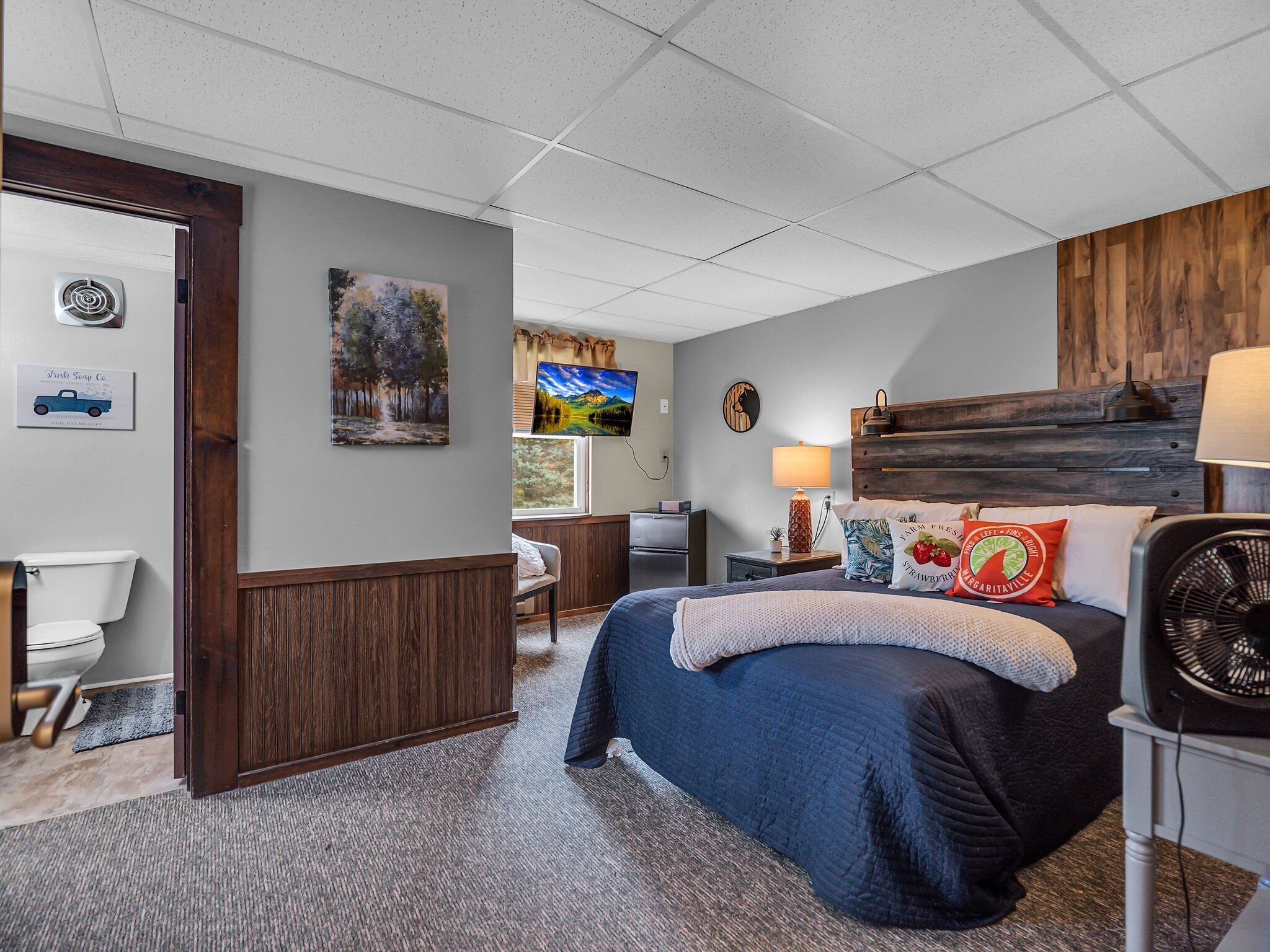 A hotel room with a bed, a desk, a chair, a TV, and a fan.
