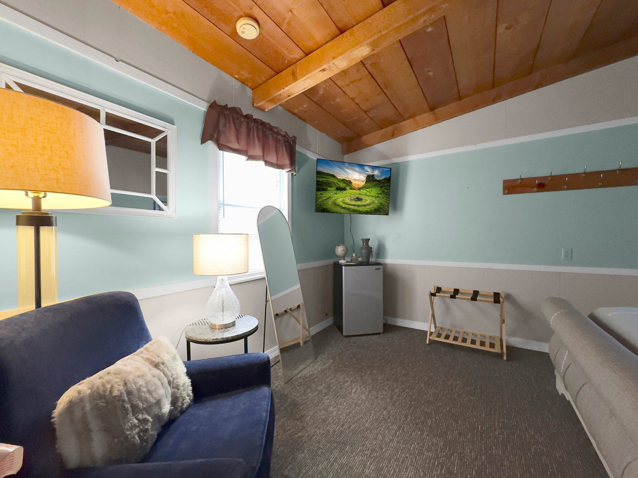 A room with a blue sofa, a lamp, a TV on the wall, and a small table with a vase.
