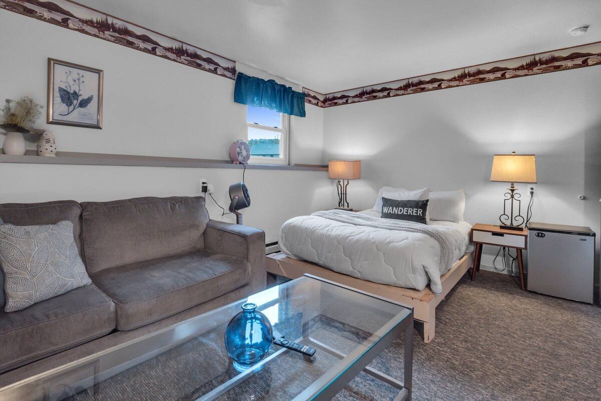 A hotel room with a bed, a sofa, a small table, a lamp, and a mini-fridge.