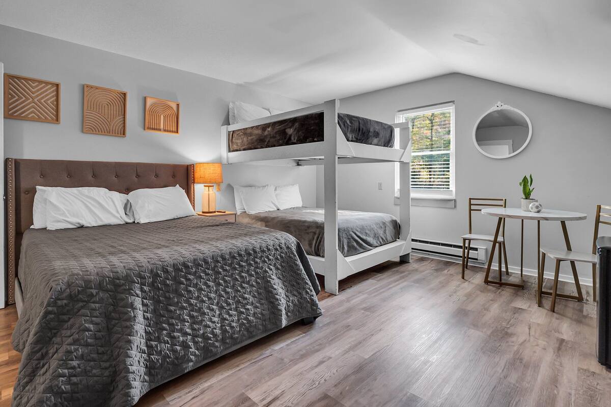 A modern hotel room with a large bed, a bunk bed, a round table with chairs, and a small potted plant.