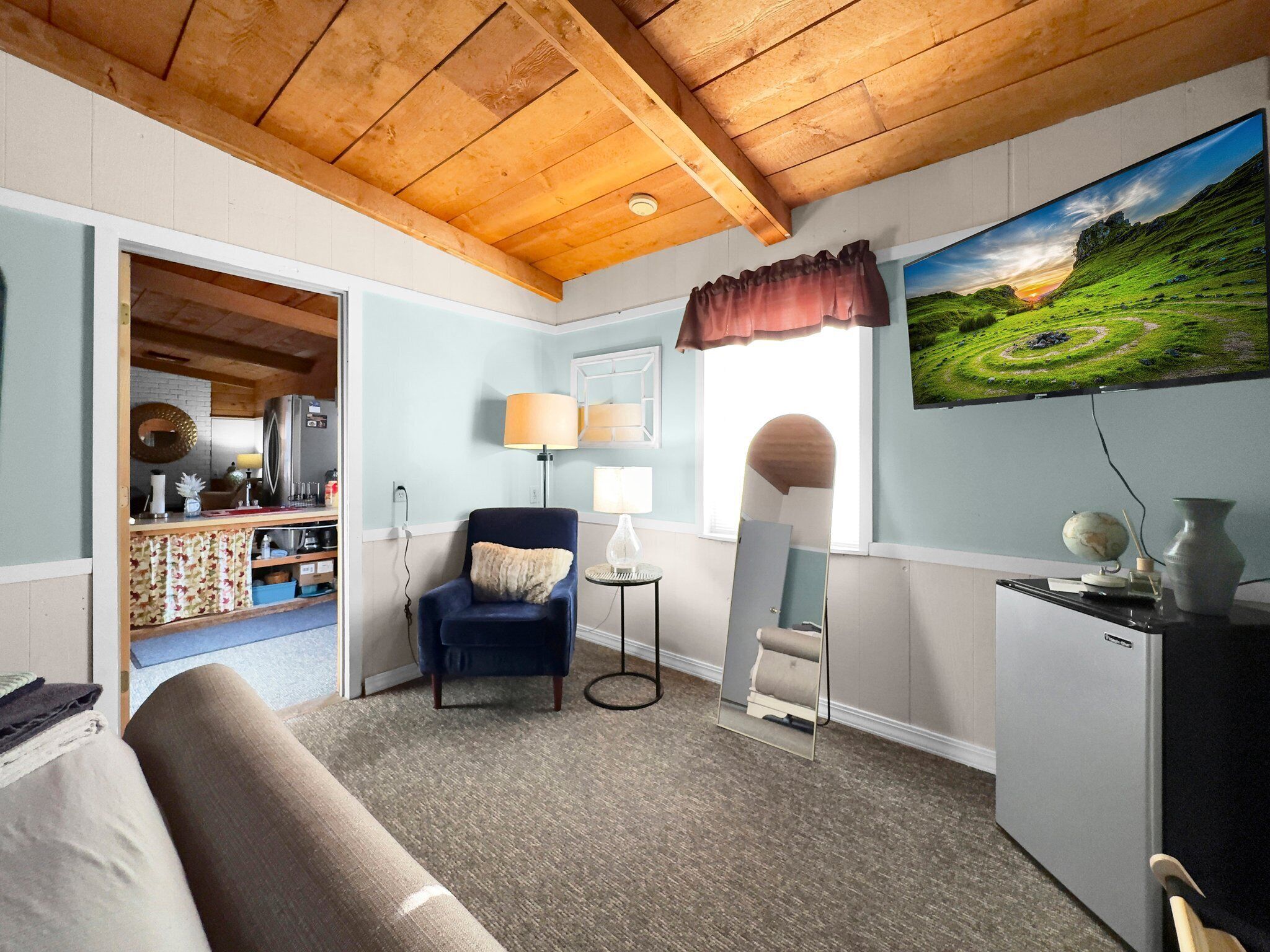 A cozy room with a wooden ceiling, a blue armchair, a small table with a lamp, a mirror, and a painting of a landscape.