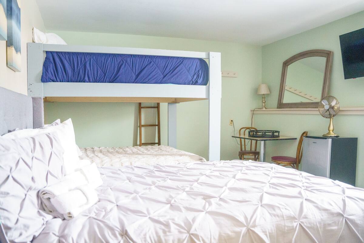 A bunk bed with a desk and chair, a small refrigerator, a mirror, and a wall-mounted fan in a room with light green walls.