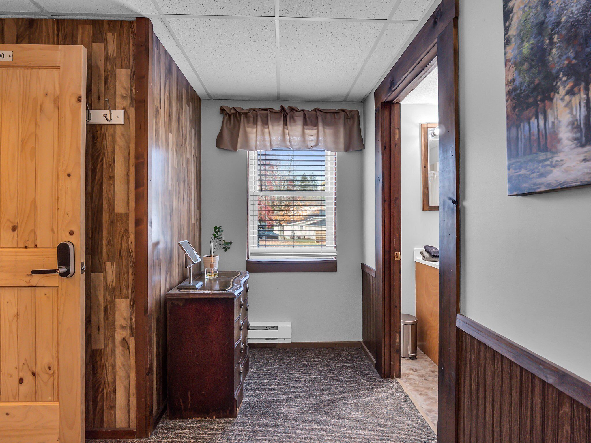 A hallway with wooden walls, a window with blinds, a desk with a plant, and a painting on the wall.