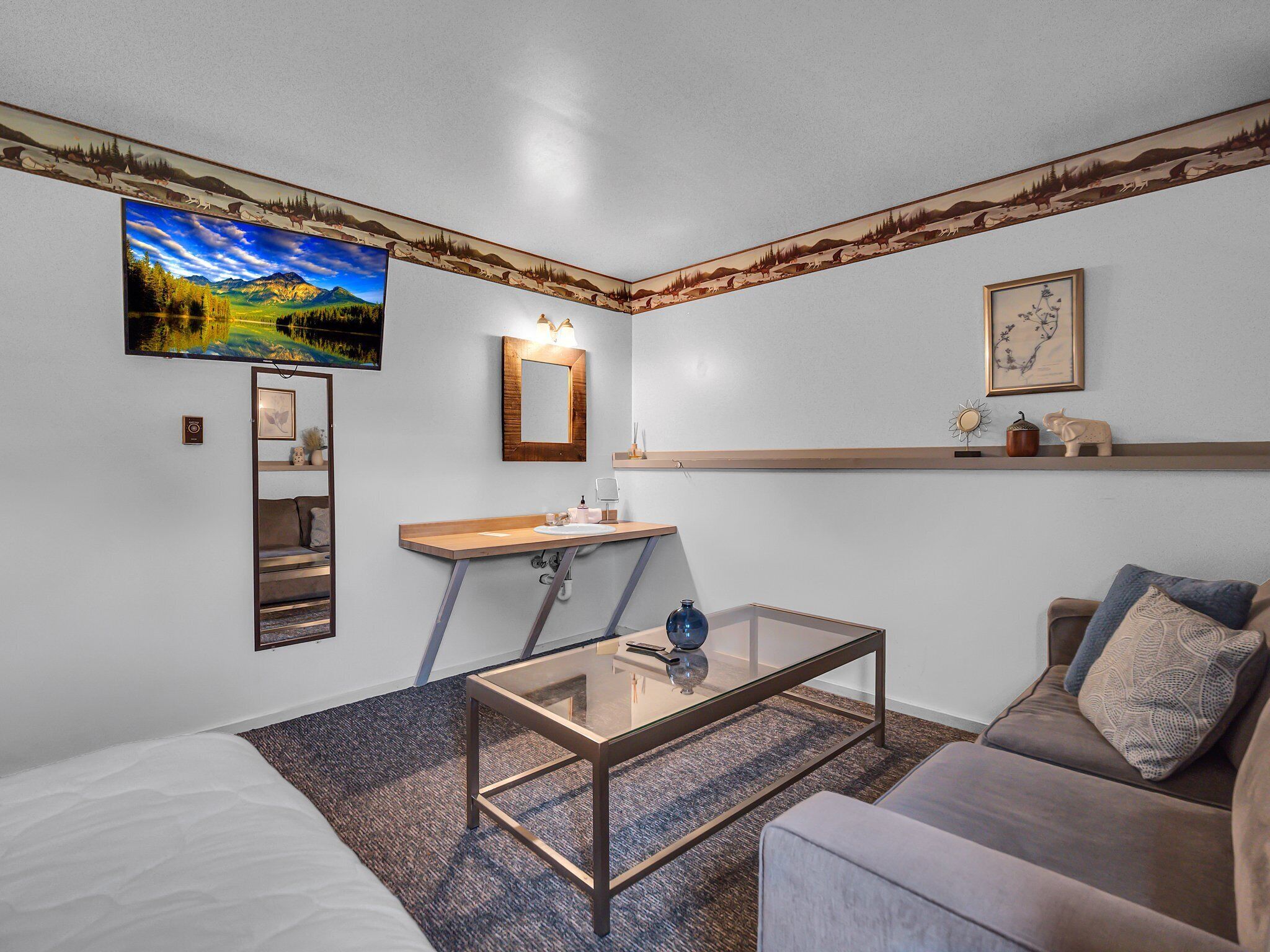 A modern room with a bed, a sofa, a glass coffee table, a small desk with a mirror, and a mounted TV.