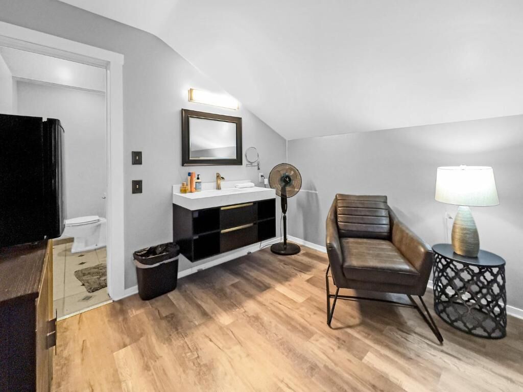 A modern living room with a white wall, a black leather sofa, a small round table with a lamp, a black floor lamp, and a white sink with a mirror above it.