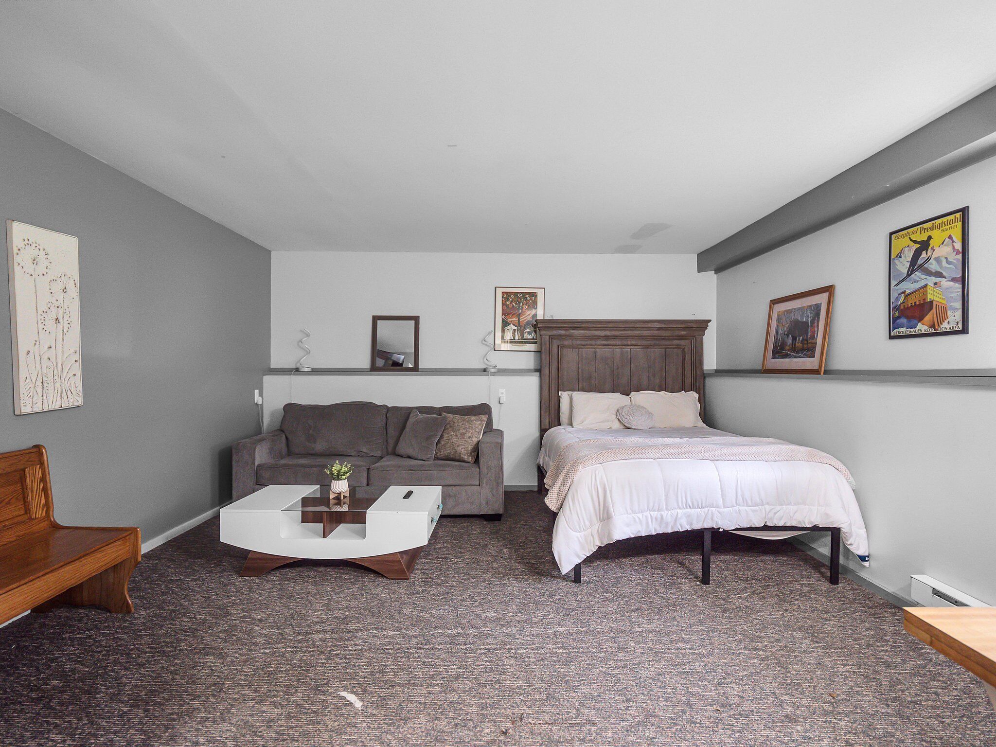 A small, modern room with a grey couch, a bed with white bedding, a wooden bench, and a coffee table.