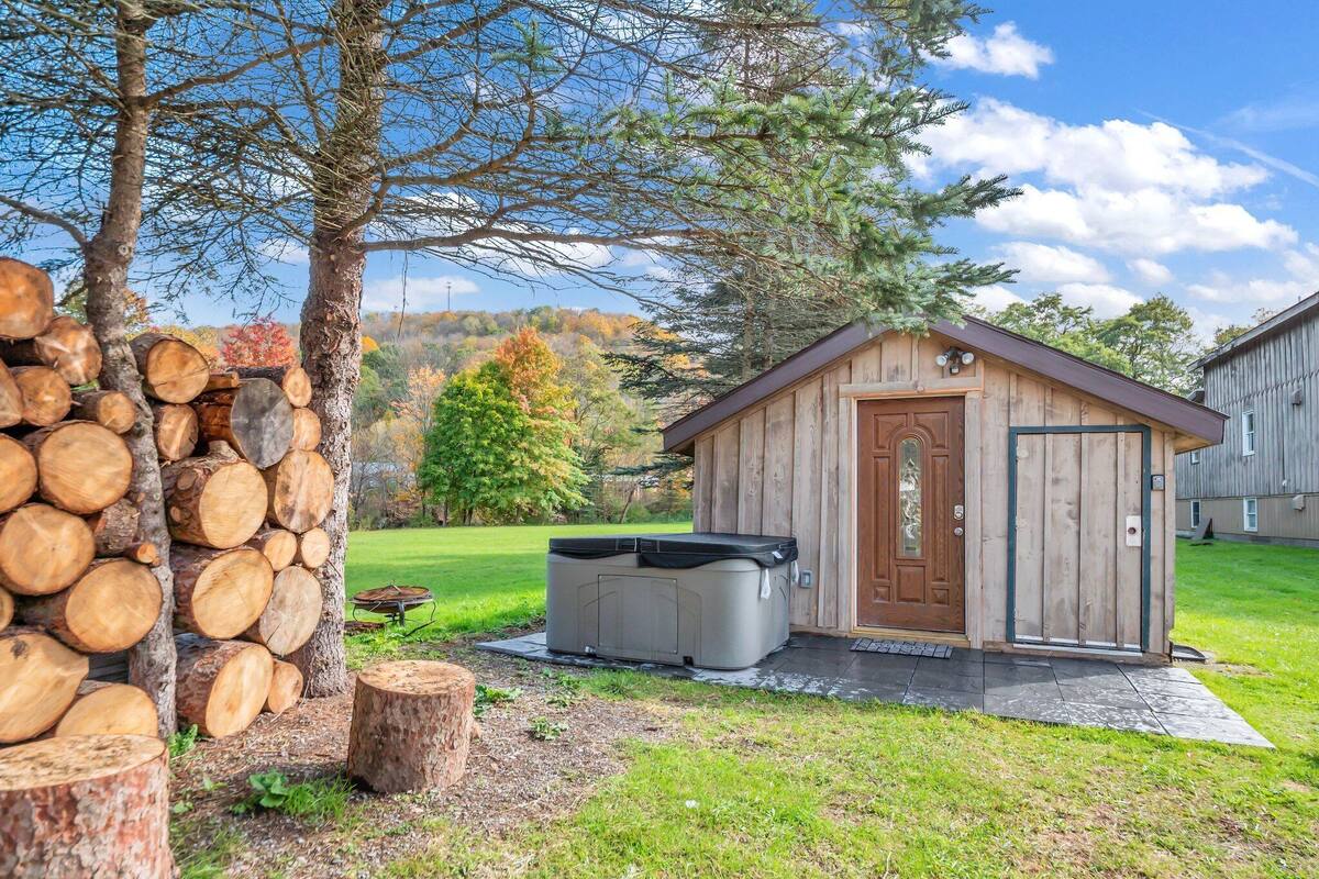 A wooden cabin with a hot tub, surrounded by a stack of logs and a tree.
