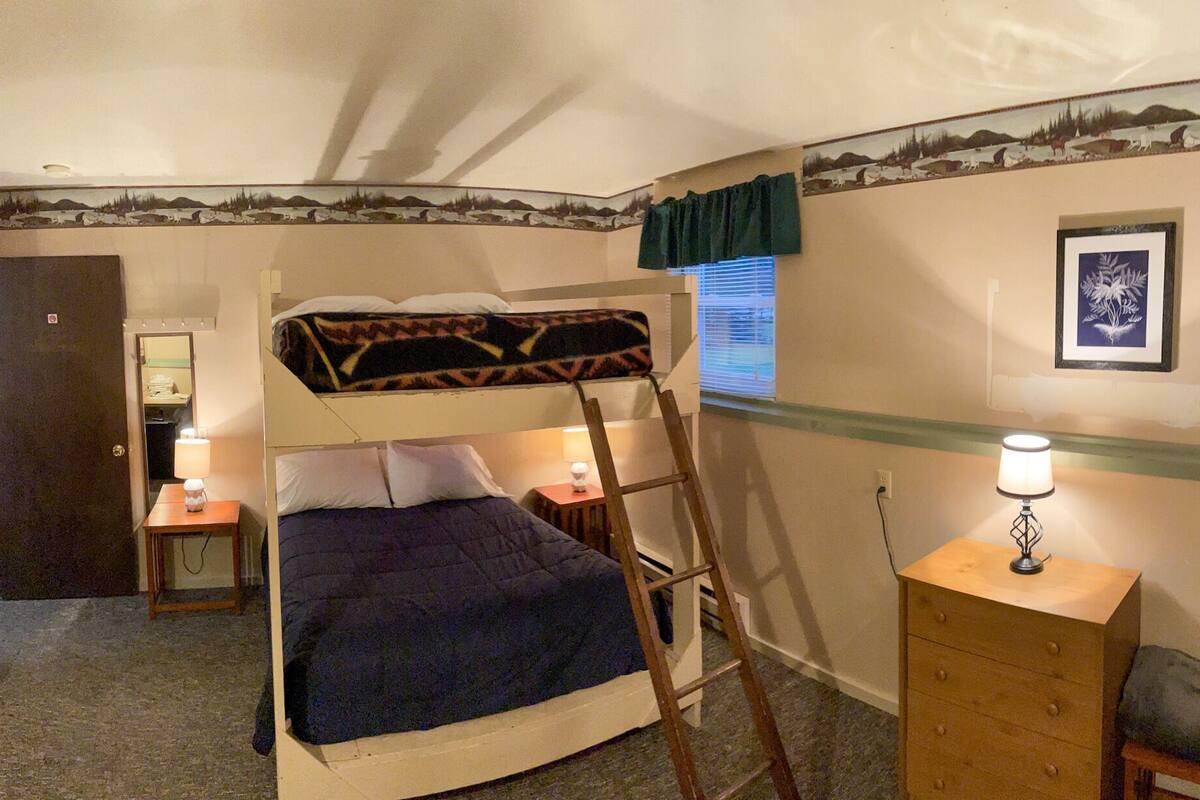 A bunk bed room with a ladder, a desk, a chair, a lamp, and a framed picture on the wall.