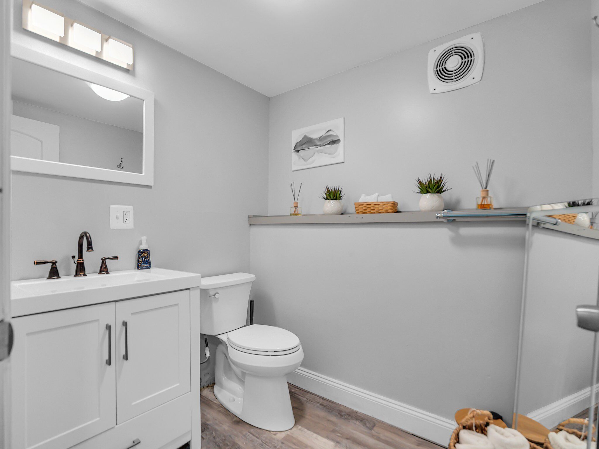Un baño moderno con lavamanos, tocador y retrete blancos. Hay un espejo con luces arriba, un ventilador montado en la pared y una repisa con plantas y una cesta.