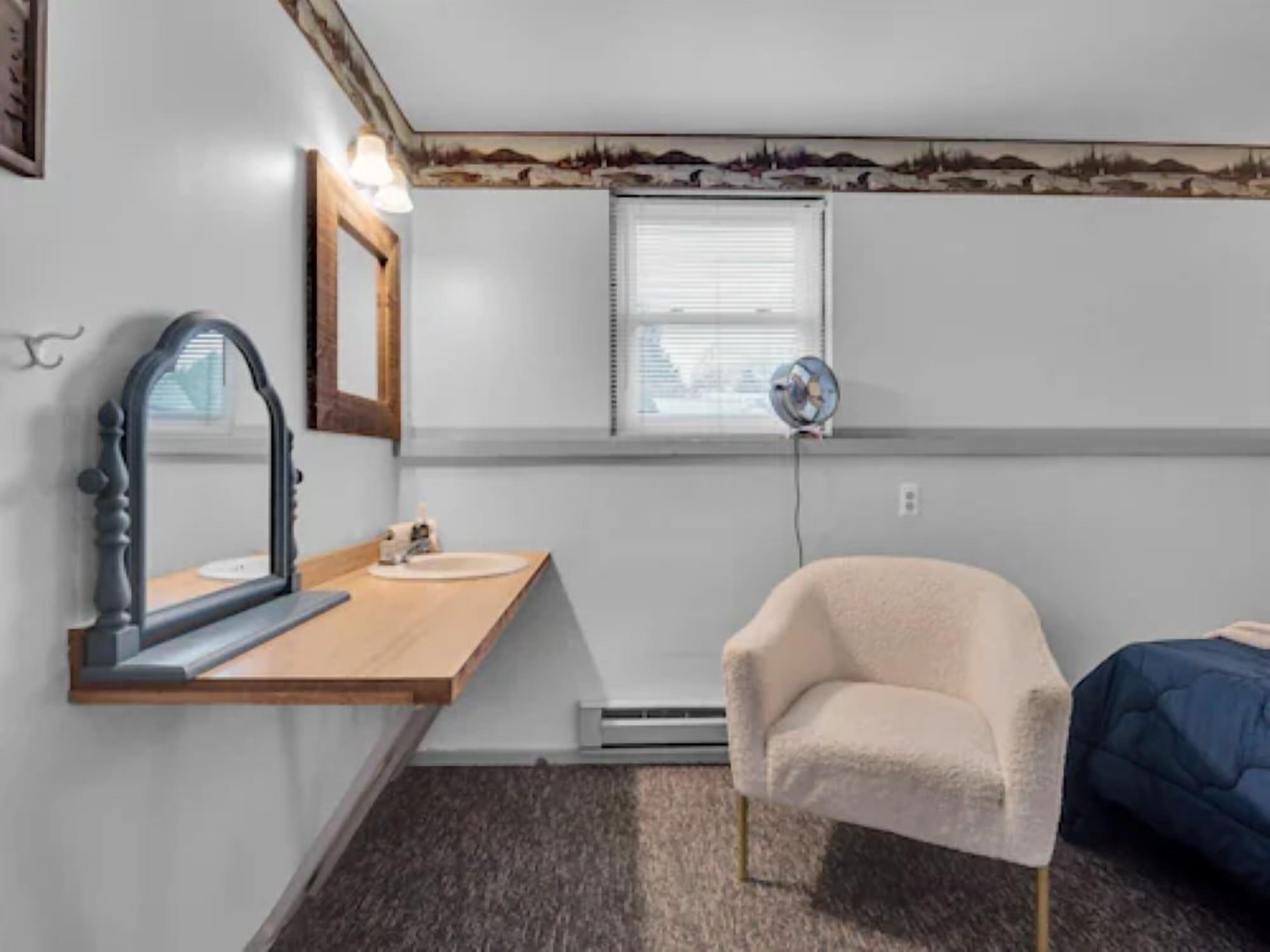 A small, compact room with a wooden vanity, a mirror, a small sink, a chair, a bed, and a window with blinds.