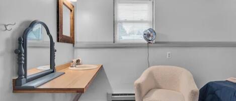 A small, compact room with a wooden vanity, a mirror, a small sink, a chair, a bed, and a window with blinds.