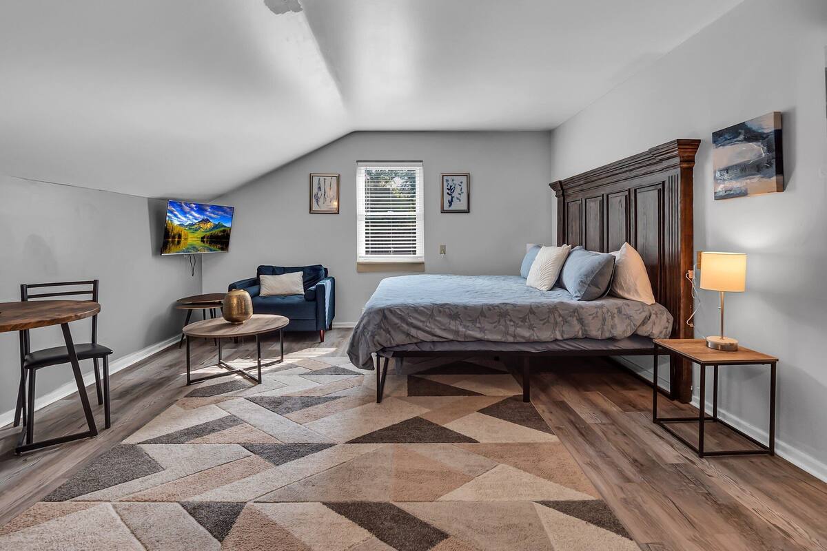A bedroom with a bed, a desk, a chair, a TV, and a window with blinds.