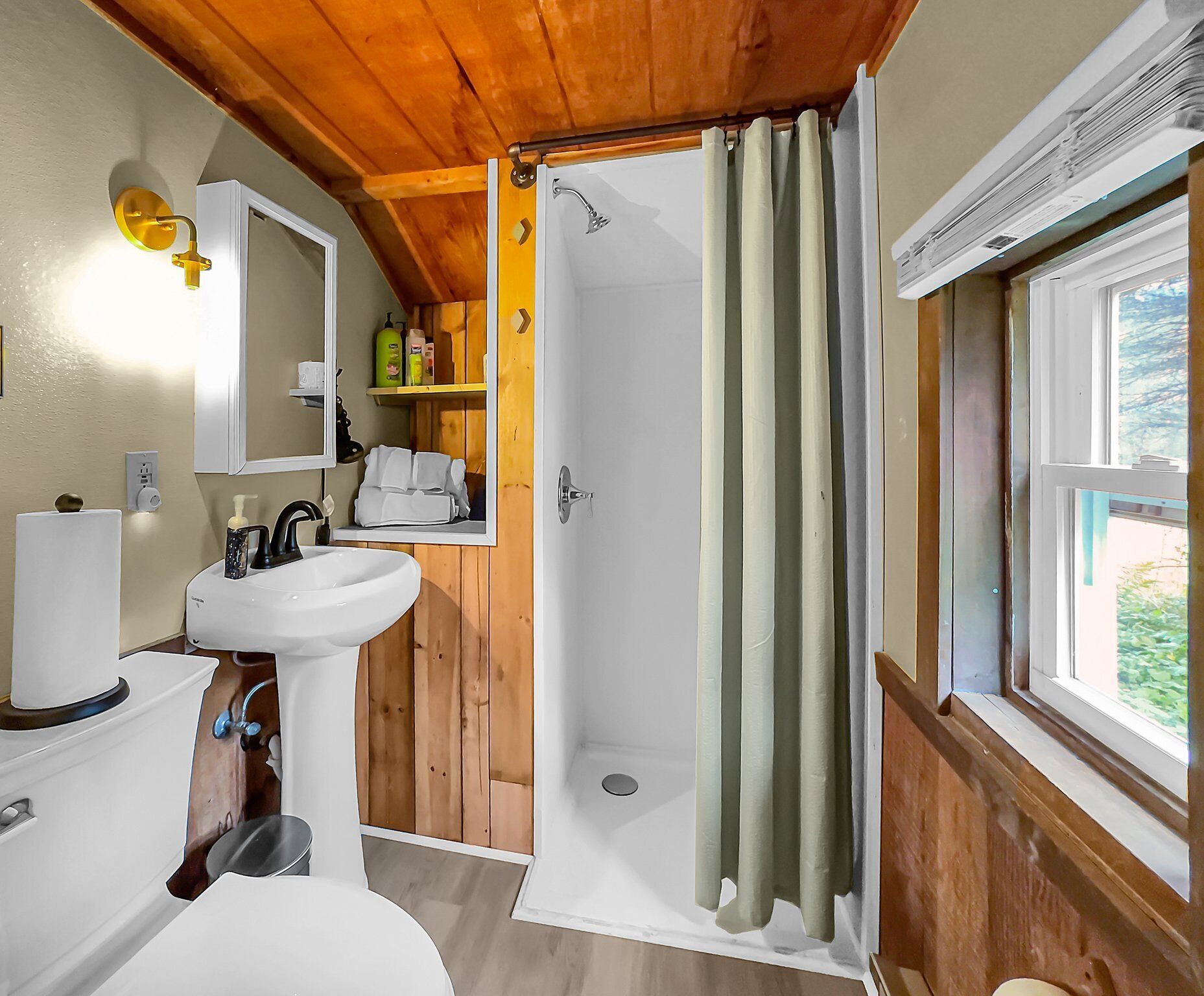 A bathroom with a wooden ceiling, a white sink, a mirror, and a shower with a green curtain.