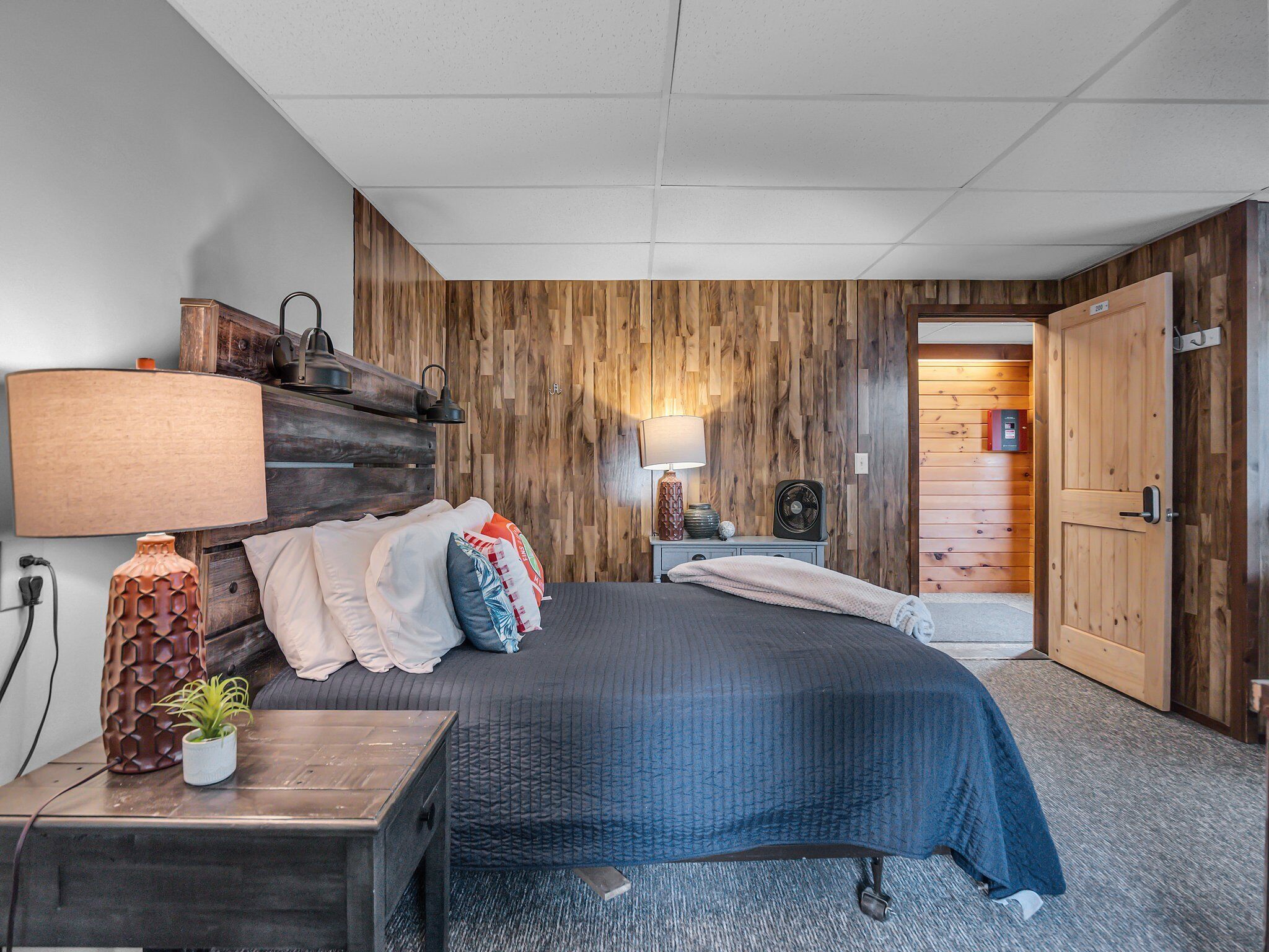 Een slaapkamer met een bed, nachtkastjes, een lamp, een ventilator en een houten wandpaneel.