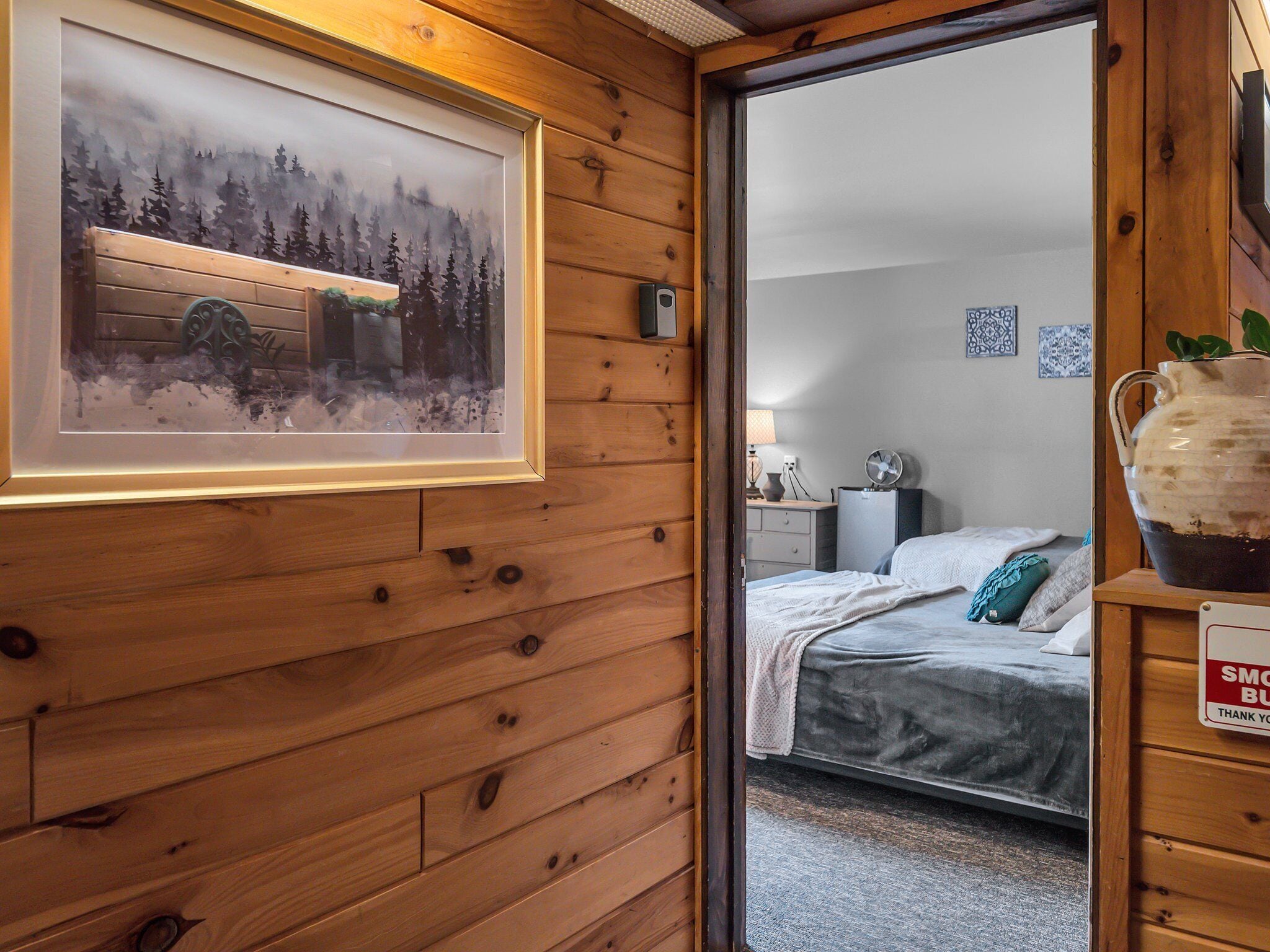 A wooden cabin interior with a framed painting of a forest, a bed with a white blanket, and a sign on the door.