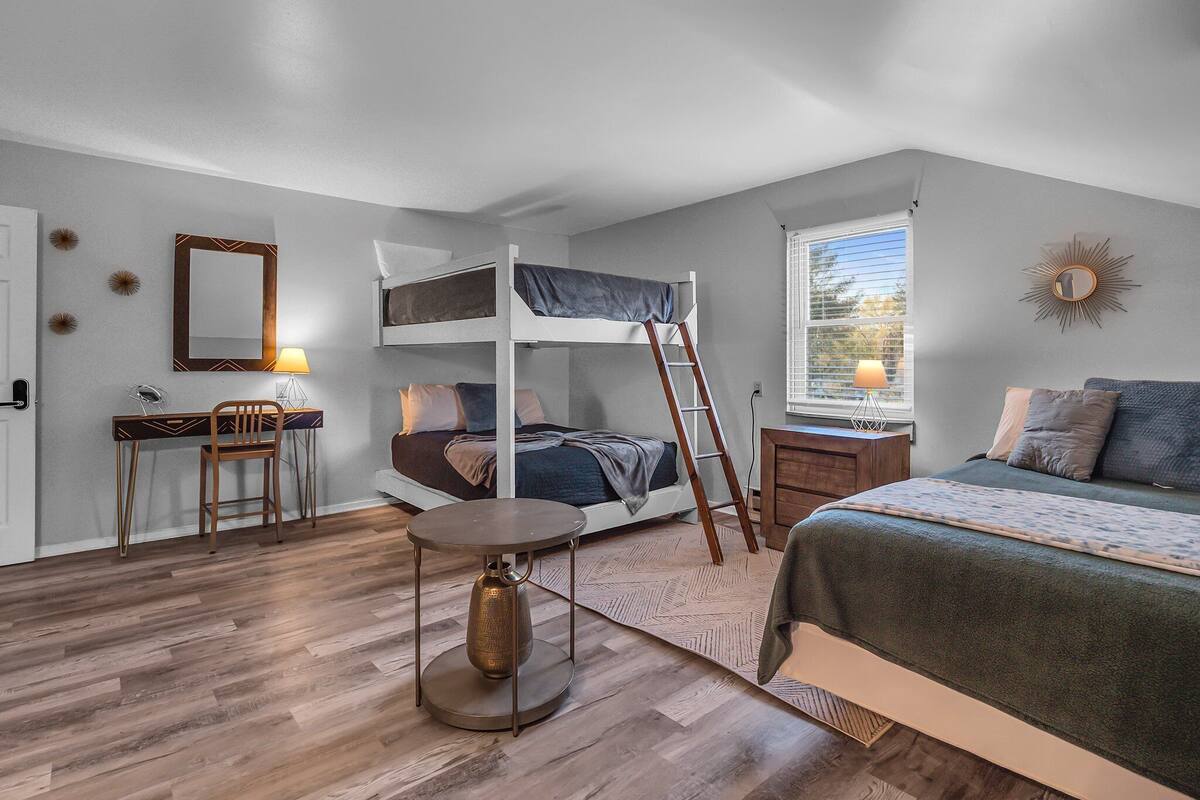 A compact bedroom with a bunk bed, a desk with a chair, a small table, and a window with blinds.