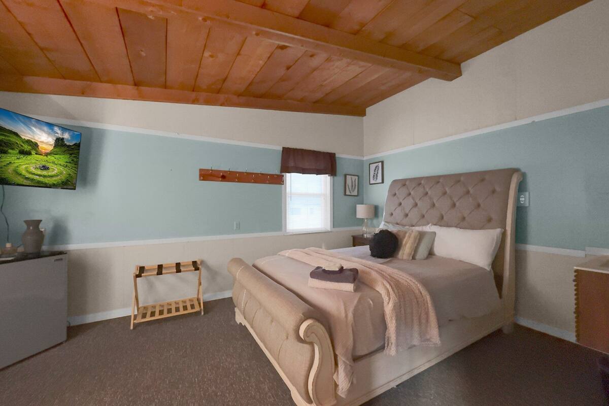 A bedroom with a bed, a wooden headboard, a small bedside table, a nightstand, a wall-mounted TV, a window with blinds, and a painting on the wall.
