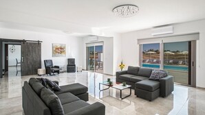 A modern living room with a glass coffee table, grey sofas, a chandelier, and a view of a pool through large sliding glass doors. - Villa Jason - Six Bedroom Villa, Sleeps 12 (Paralimni)