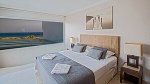 Une chambre avec une grande fenêtre donnant sur une plage, un lit avec une literie grise et un tableau accroché au mur.