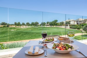 Spisebord med udsigt til en golfbane, en glasafskærmning og et måltid med tallerkener, bestik og glas.