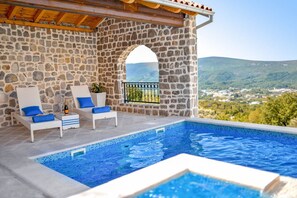 Une piscine à débordement aux murs de pierre, agrémentée de deux chaises longues blanches et de coussins bleus, offrant une vue imprenable sur un paysage magnifique.