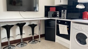 A compact kitchen with a flat-screen TV, a microwave, a small refrigerator, a washing machine, and three bar stools. - Modern 2 Bedroom Flat in Robert st, Swansea (Swansea)