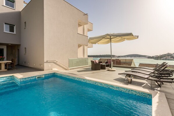 A rooftop pool with clear blue water, a seating area with lounge chairs and an umbrella, and a view of the sea and distant hills.