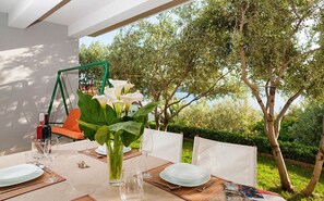 An outdoor dining area with a table set for two, surrounded by greenery and a view of trees and water.