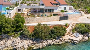 Una propiedad costera con piscina, escalones de piedra que bajan a una playa y una casa moderna con tejado de tejas rojas.