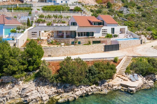 Une propriété située sur la côte, dotée d’une piscine, d’un escalier en pierre menant à la plage et d’une maison moderne avec un toit recouvert de tuiles rouges.