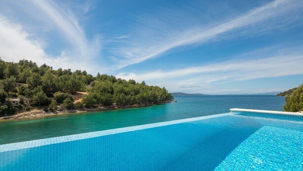 Una piscina infinita azul cristalina con vistas al mar tranquilo y a una ladera boscosa.