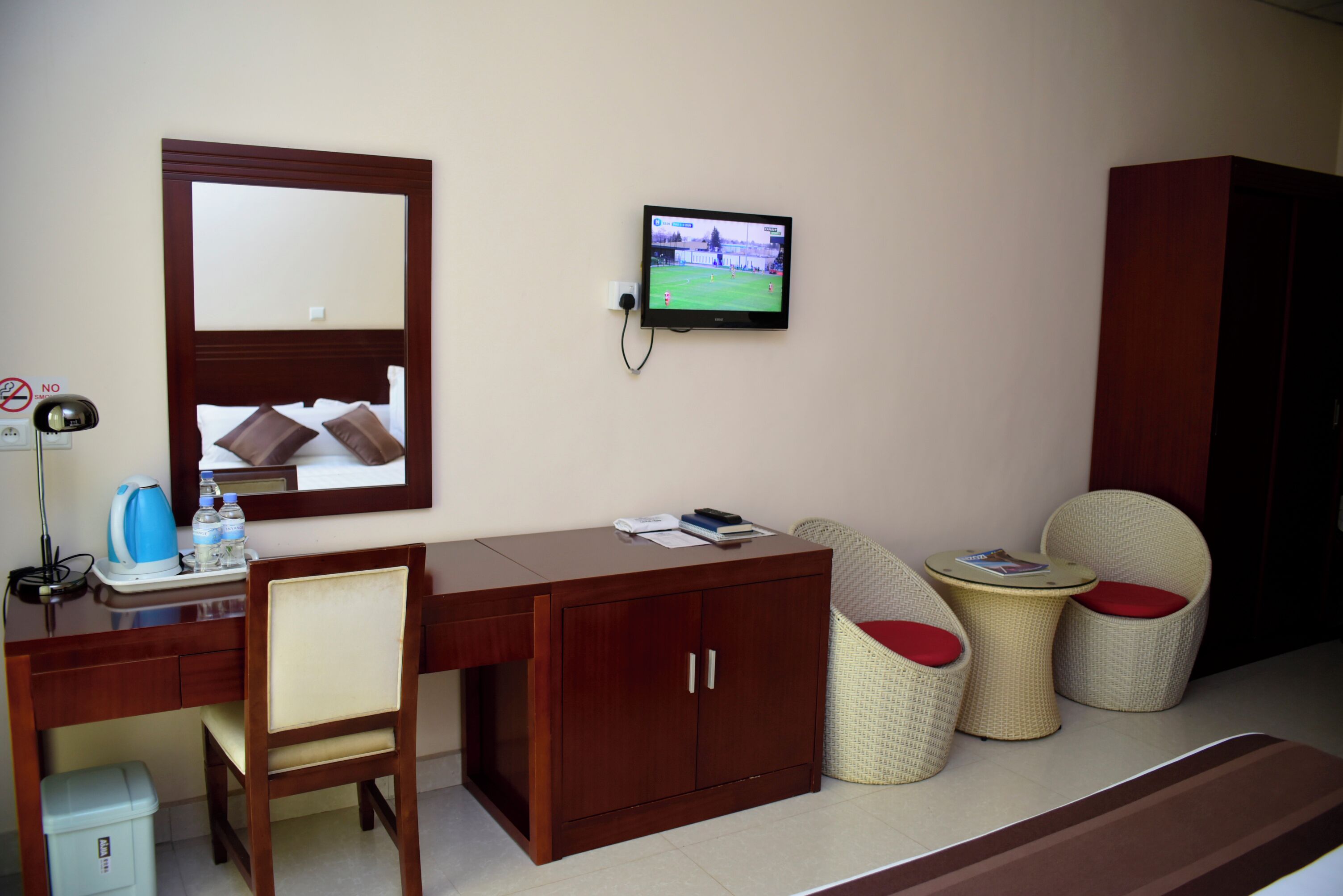 A hotel room with a wooden desk, a television mounted on the wall, a chair with a red cushion, a small round table, and a wardrobe.