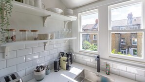 Uma cozinha com azulejos brancos, uma pia, uma janela com vista para edifícios e diversos utensílios de cozinha nas prateleiras.