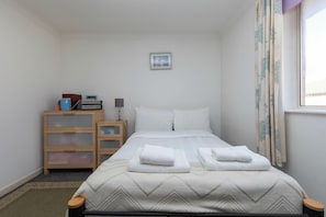 Habitación de hotel ordenada con una cama, una mesita de noche con lámpara, un armario pequeño y un ventanal con cortinas.
