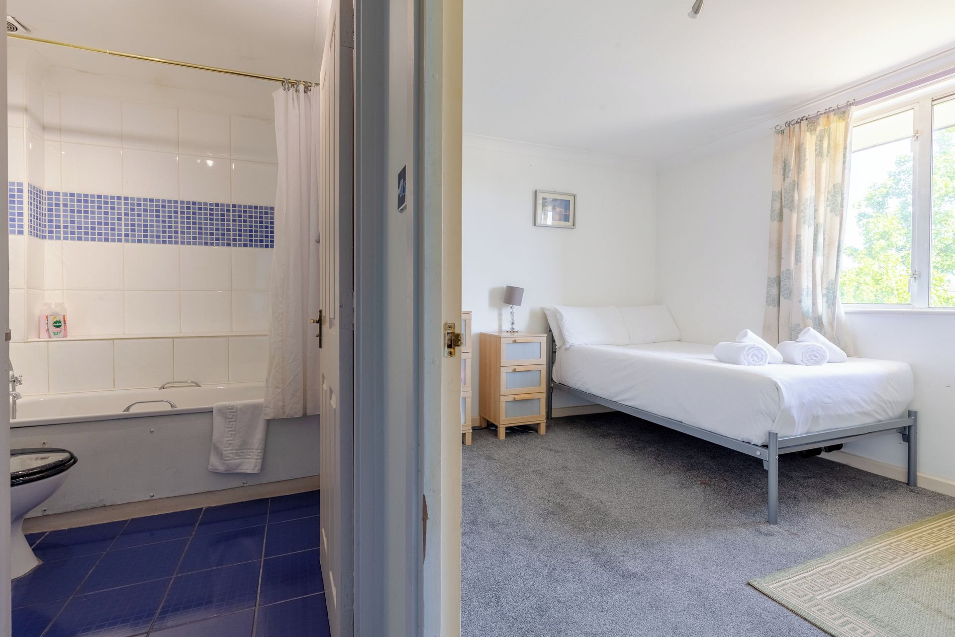 Un dormitorio con cama individual, mesita de noche, ventana con cortinas y baño con bañera e inodoro.