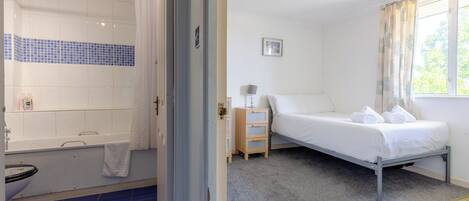 Un dormitorio con cama individual, mesita de noche, ventana con cortinas y baño con bañera e inodoro.
