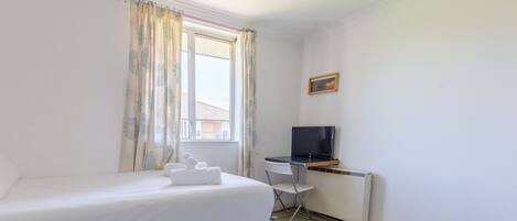 A small, clean room with a single bed, a desk with a computer, and a window with curtains.
