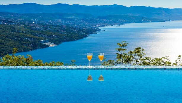 Ein Fernblick über eine Küstenstadt und Berge auf einen Infinity-Pool mit zwei Orangensäften.