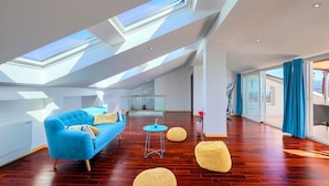Ein großzügiger Wohnbereich mit hoher Decke, Oberlichtern, einem blauen Sofa, Holzboden und einer Glasdoor, die zu einem Außenbereich führt.