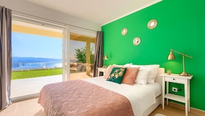 Ein Schlafzimmer mit grüner Wand, einem großen Bett, einem Nachttisch und Blick ins Freie.