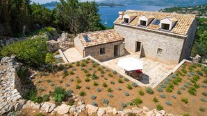 A stone building with a tiled roof, a small garden with shrubs, and a view of the sea and mountains in the background.