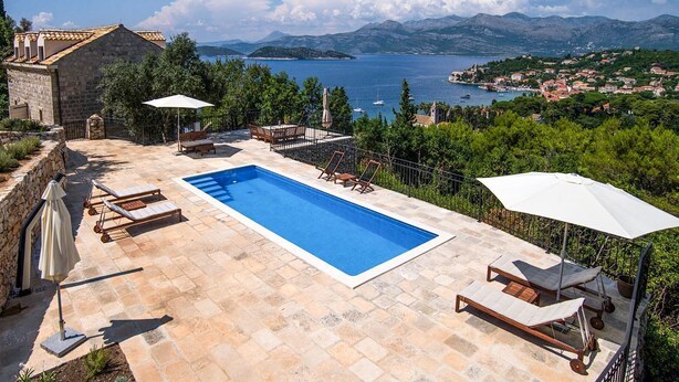 A swimming pool with lounge chairs and umbrellas, surrounded by stone walls and a view of the sea and mountains.
