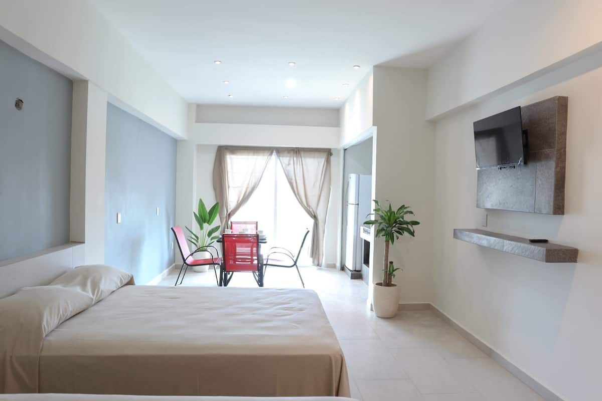 Une chambre d’hôtel moderne avec un grand lit, un coin repas avec des chaises rouges, une plante en pot et un téléviseur fixé au mur.