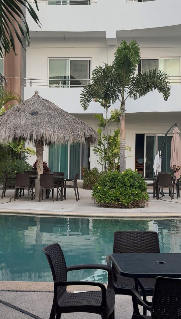Un área junto a la piscina con una sombrilla de paja, sillas de comedor y una pileta.
