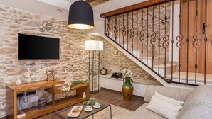 Un salon avec un mur en pierre, un téléviseur à écran plat, une console en bois, une table basse avec des pâtisseries, un canapé et un escalier avec des rampes en fer forgé.