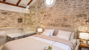 Une chambre avec un mur en pierre, un grand lit, une baignoire et une lucarne ronde.