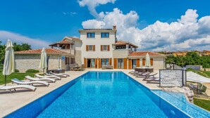 Vue sur une piscine d’hôtel, avec une grande piscine, des chaises longues et un bâtiment à deux étages surmonté d’un toit en tuiles.