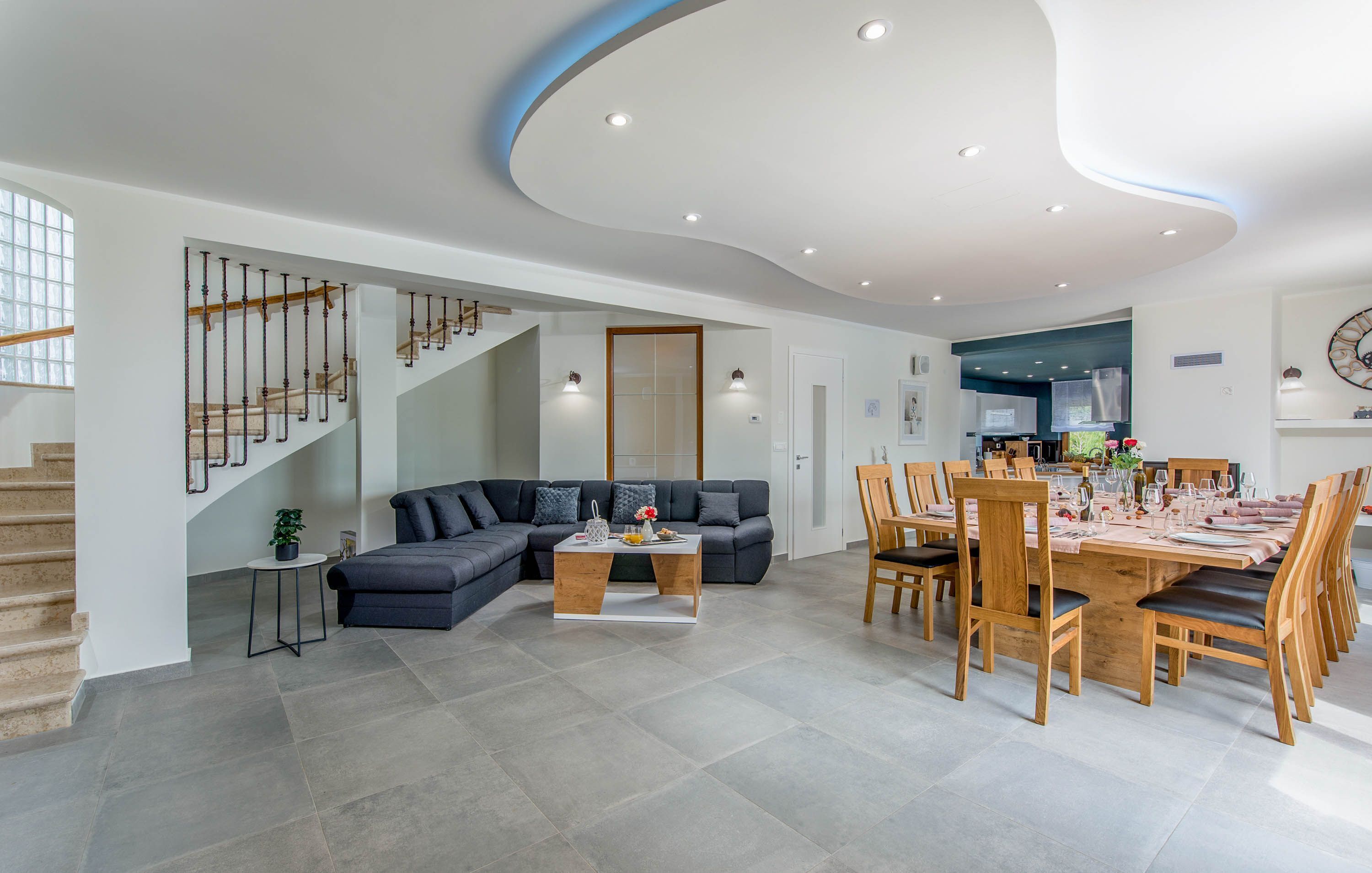 A spacious living area with a dark-colored sofa, a wooden coffee table, and a dining table set for six. A staircase with wooden railings leads to an upper floor.