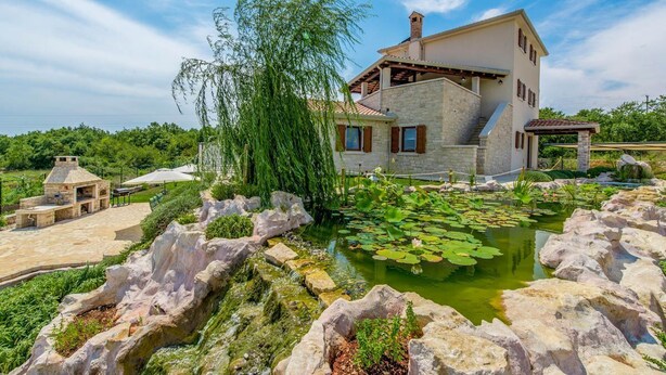 Une maison aux murs en pierre, un étang parsemé de nénuphars et un saule pleureur.